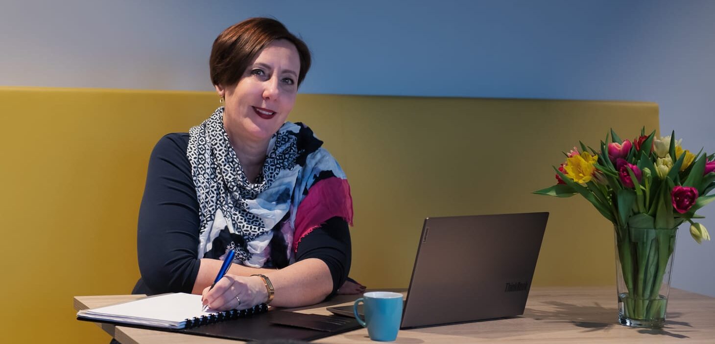 Home Carola zit op een gele bank schrijvend in een map met een laptop op de tafel en een bos tulpen